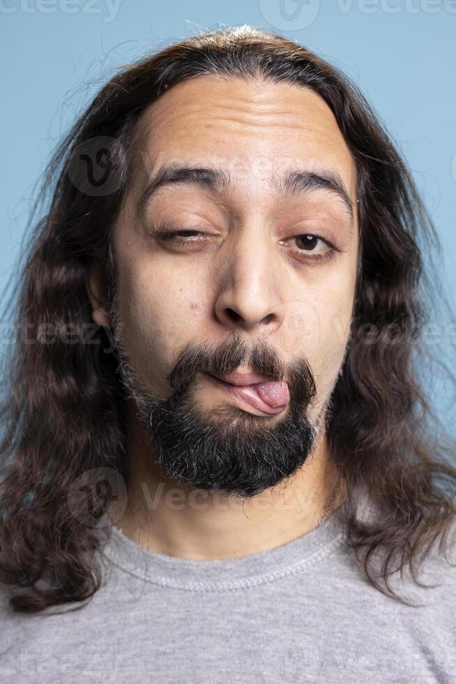 Cheerful overjoyed guy with long hair and beard making a hilarious grimace, fooling around on camera. Humorous relaxed man brings a sense of joy and laughter to the blue backdrop. photo
