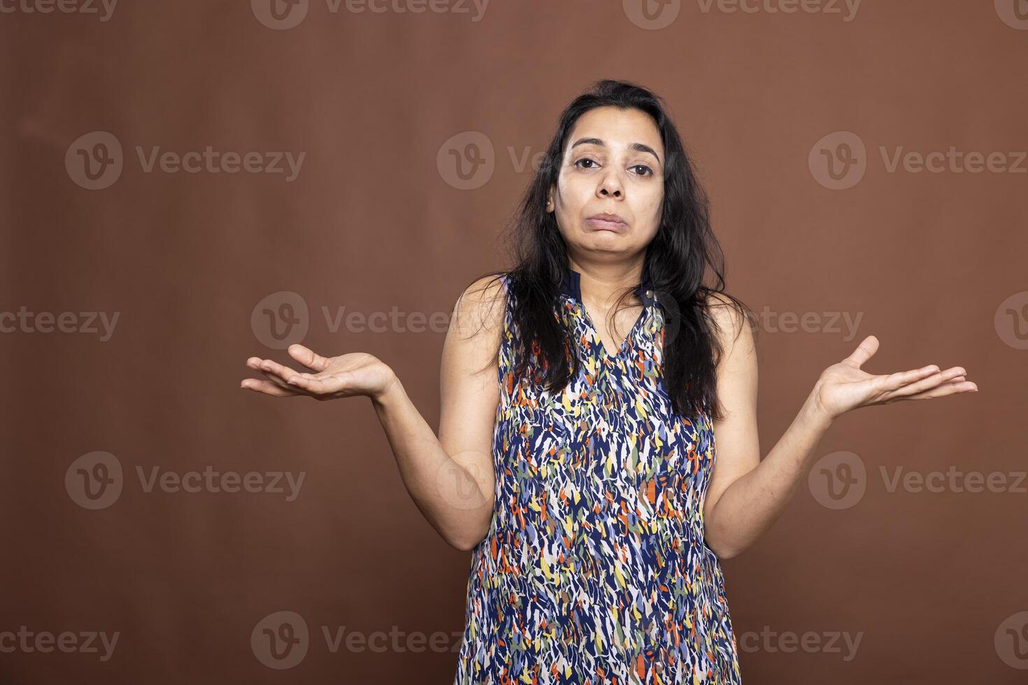 Confused indian lady with pursed lips and shrugged shoulders, expressing uncertainty and ignorance. Pretty female individual standing and looking unsure against isolated background. photo