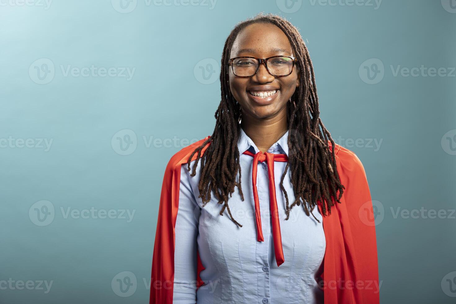 Portrait of black female individual cheerfully standing isolated over blue background. Smiling young lady with glasses and red superhero cape, looking at camera, portraying positivity in modern studio photo