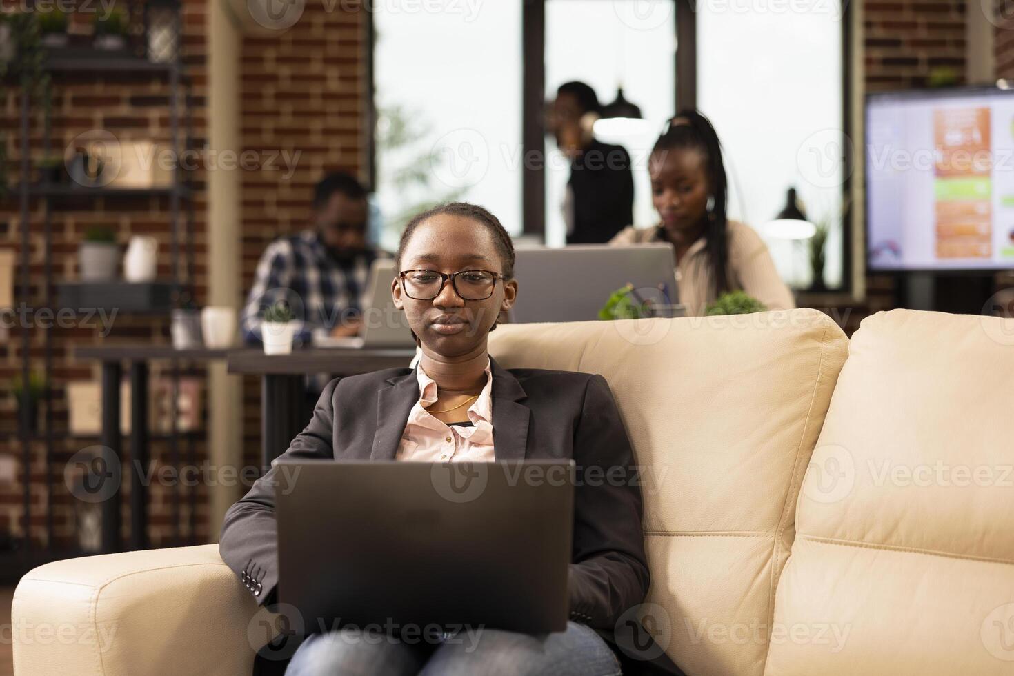 Black female manager sits on cozy couch, using laptop to analyze startup research data and manage company reports. Focused businesswoman with glasses, reviewing emails and preparing business projects. photo