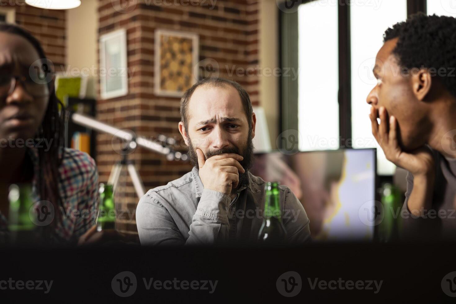 Concerned white man watching emotional content with african american friends in brick wall room. Bearded male individual sitting at table, appears distressed while viewing sad with covered mouth photo