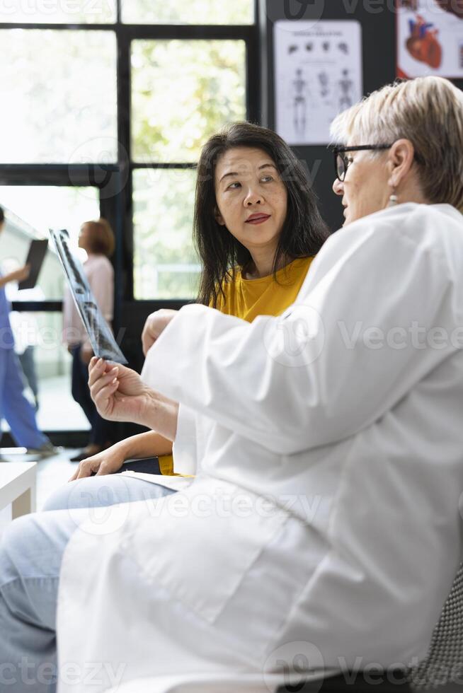 Inside a hospital lobby, medic does radiography scan interpretation with asian woman during checkup. Diverse people discussing about health evaluation and treatment guidance. Lifestyle awareness. photo