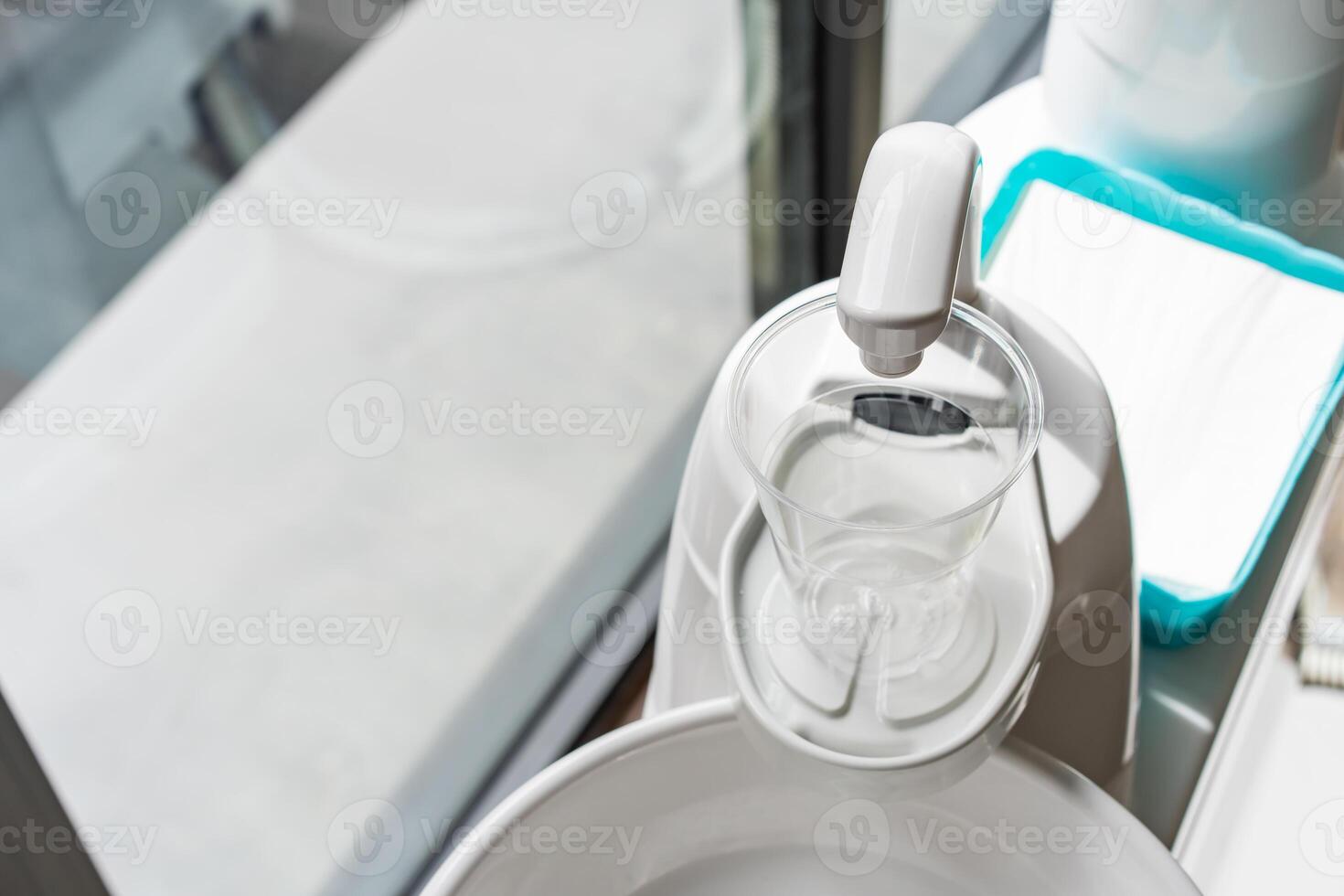A plastic glass on a cup holder of the modern ceramic mouth cleaning sink in the dentist office near a window,a photo