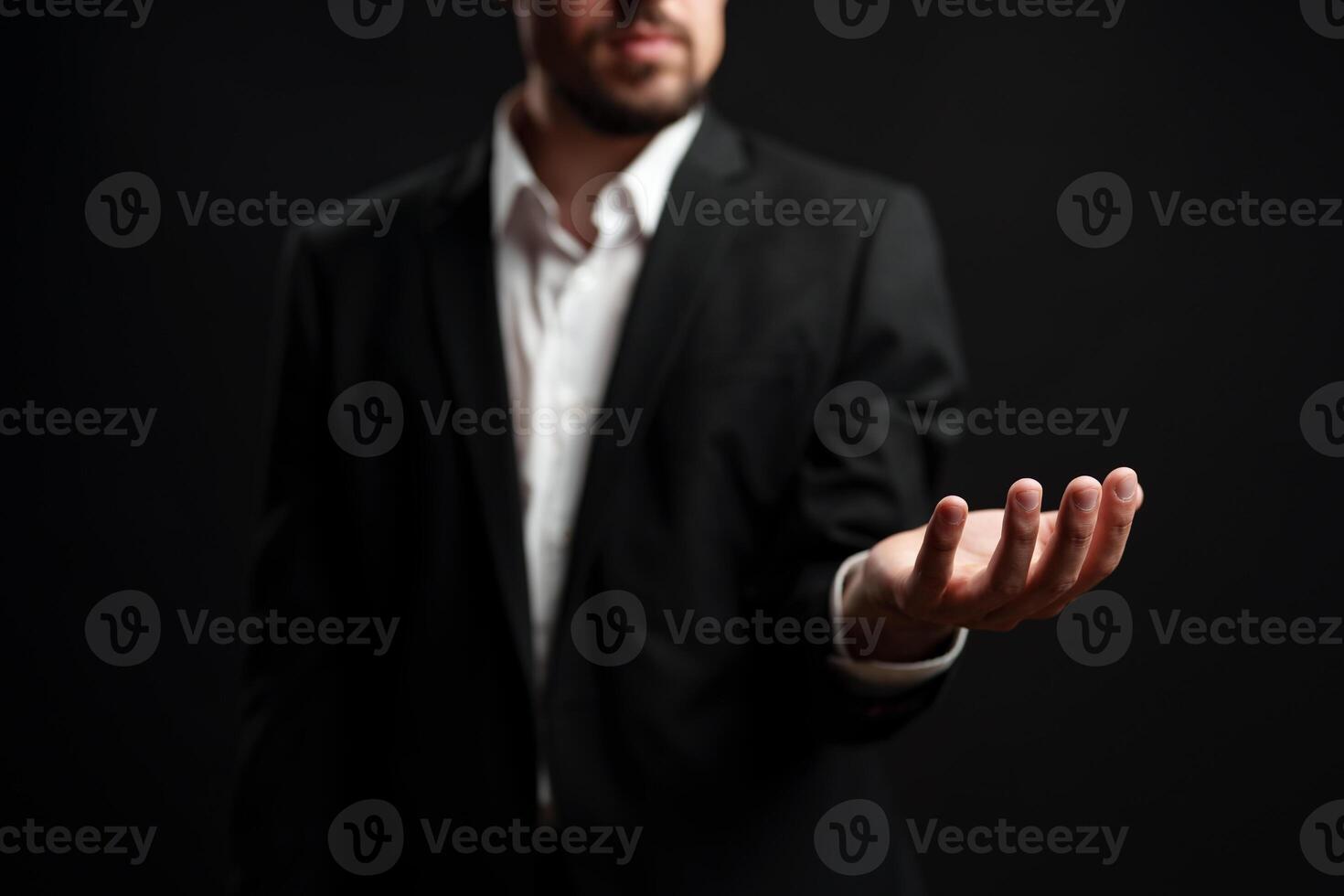 Businessman in formal attire extends hand in welcoming gesture against dark background during networking event photo