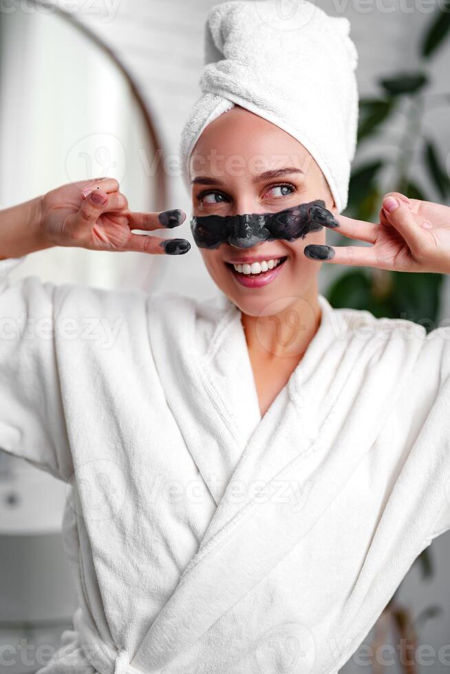 Woman applying black facial mask while smiling in a bathroom wearing a white robe and towel photo