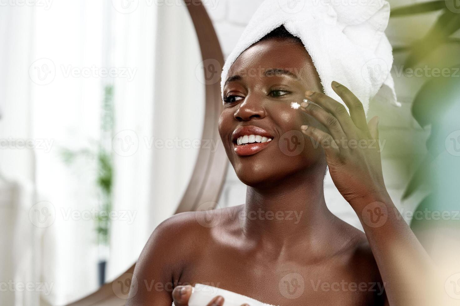 Woman applying moisturizer in a bright bathroom with a towel on her head during skincare routine photo
