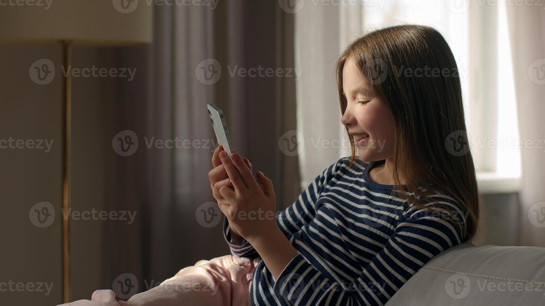 Caucasian happy little girl lying on sofa holding mobile phone smartphone cellphone gadget child kid daughter technology texting chat online pleasure smile home connect internet sms communicate female photo