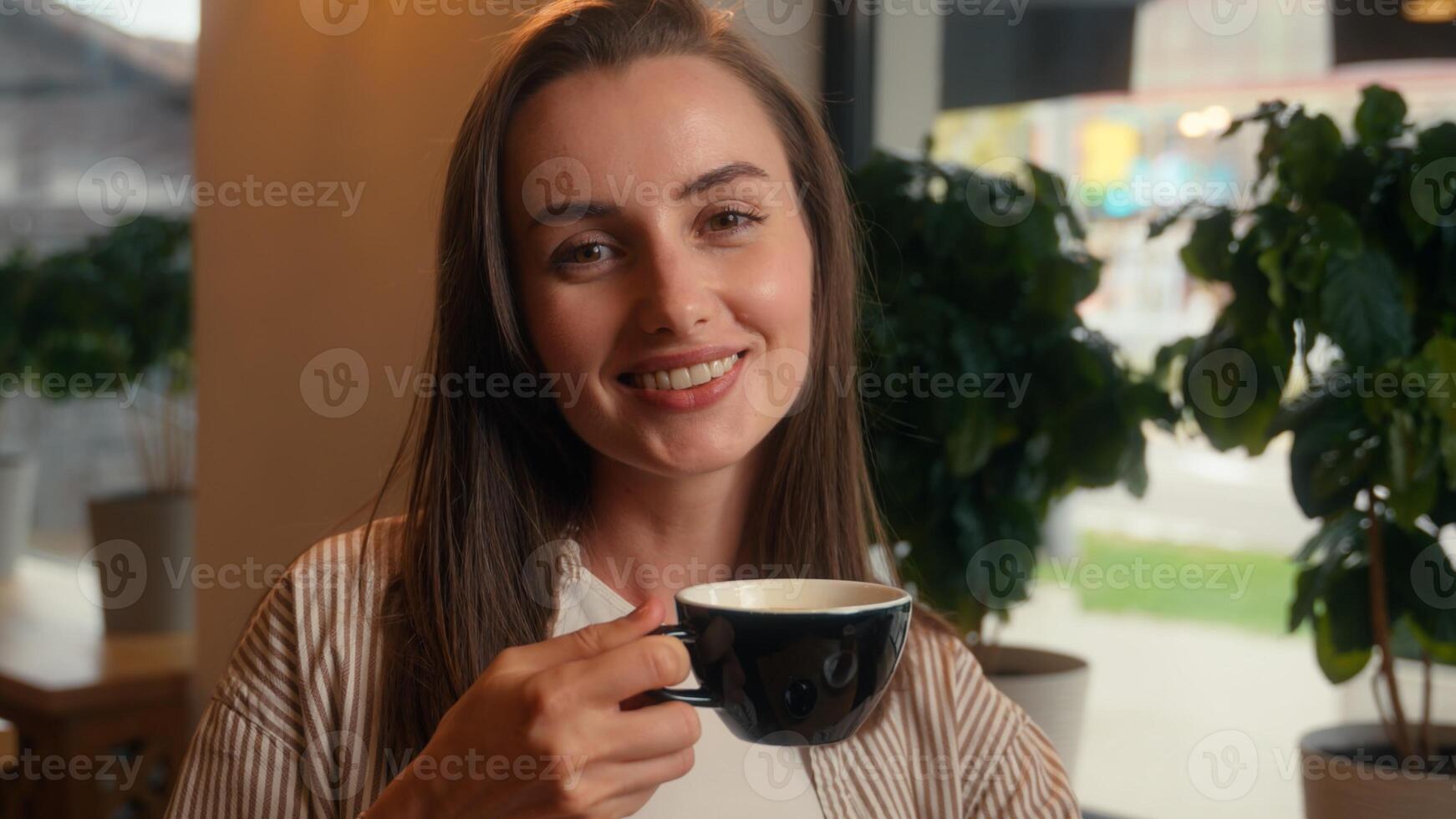 Relaxed Caucasian woman drinking coffee in cafe girl female sitting at table in cafeteria visitor drink tea croissant morning breakfast pause in restaurant looking at camera smiling happy satisfied photo