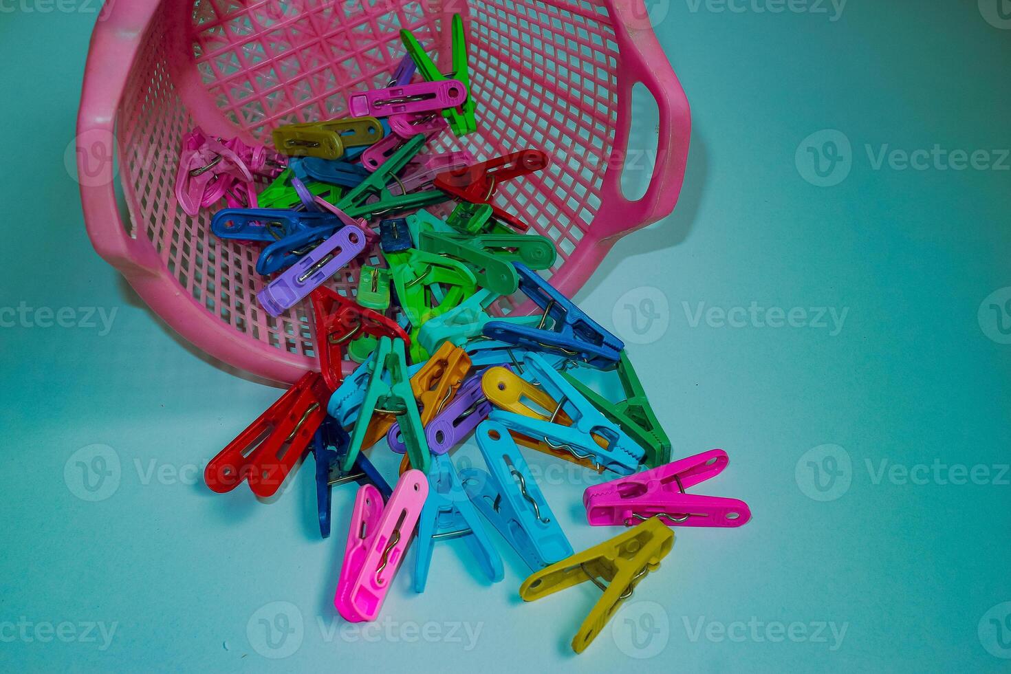 Playful design Colorful plastic clothespins scattered from pink basket on blue-green background. Playful everyday household objects in bright, abstract pop art flat lay. photo