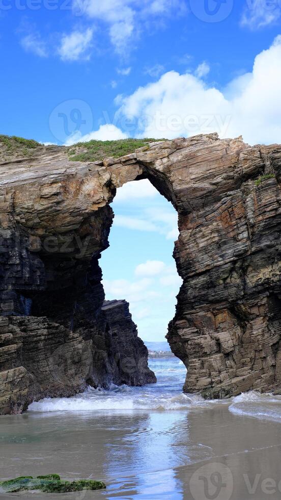 A stunning natural arch carved into towering sea cliffs on a sandy beach, with the ocean visible through the opening. photo