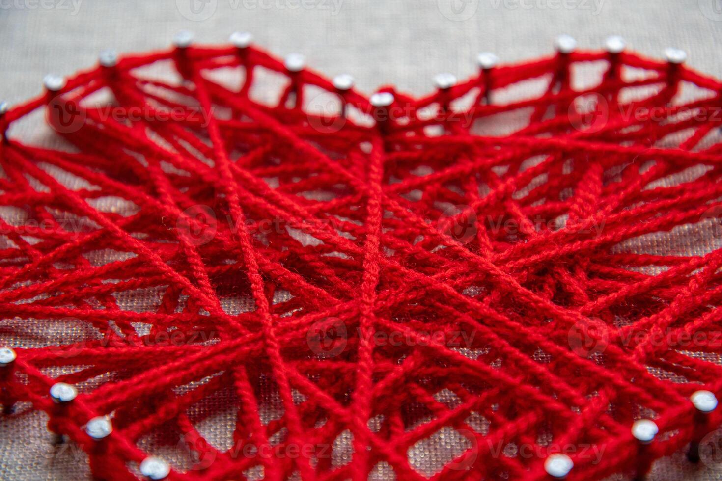 Colorful and intricate string art displays a heart shape formed by intertwined red yarn wrapped around small nails on a textured background. photo