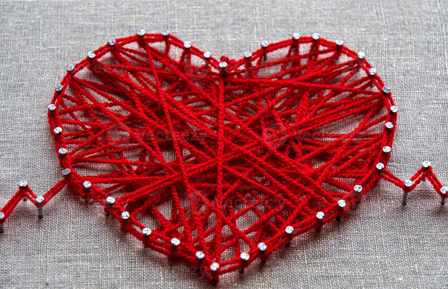 Craft string art in shape of heart. Red woolen heart, symbol of love, made of red wool yarn threads tangled over metal nails on canvas background photo