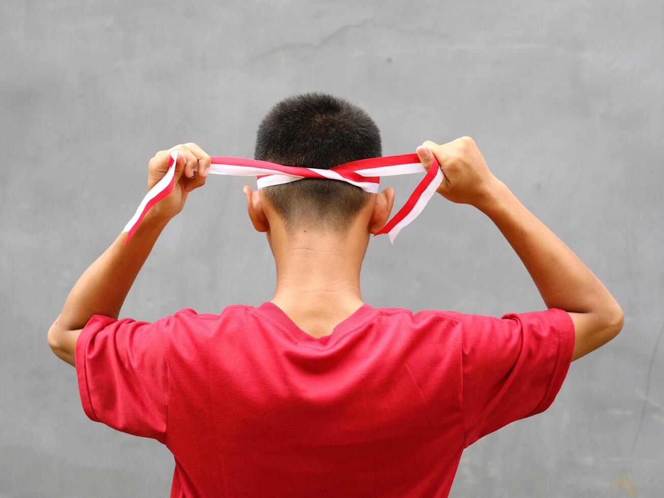 Indonesia boy in red shirt with Indonesian flag on his shoulder, his hand holding ribbon on his head, back view, Indonesia independence day celebration concept photo