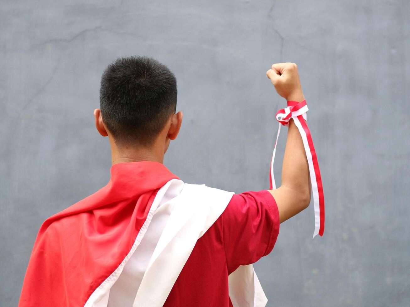 Young boy in red shirt with Indonesian flag on his shoulder, his fist raised upwards, during Indonesia's Independence day celebration, back view photo