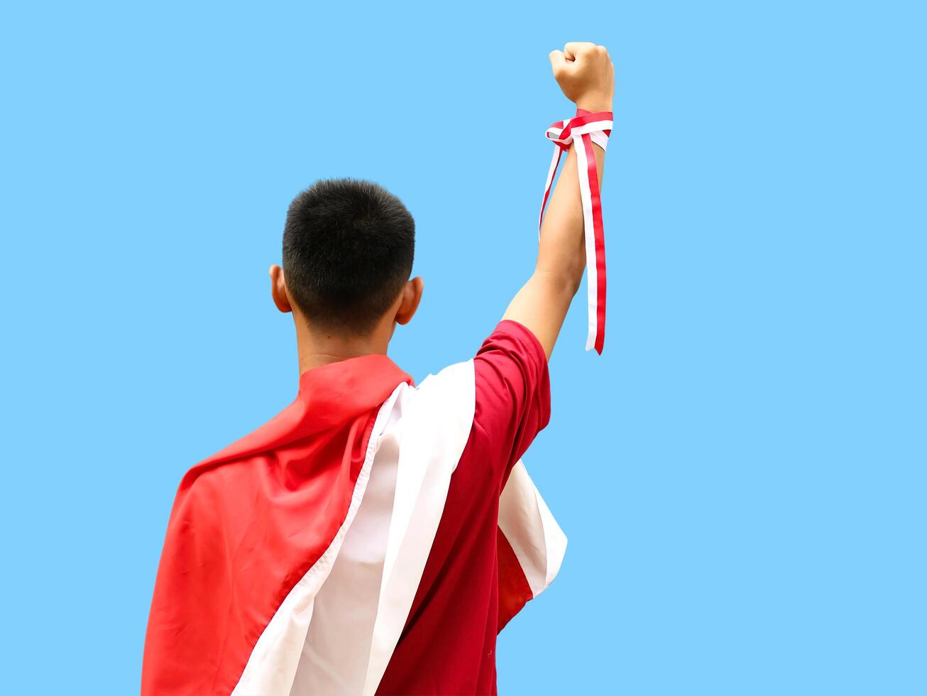 Indonesian boy in red shirt with red and white ribbon on his head, raising hand up, back view, blue background, Indonesia independence day celebration concept photo