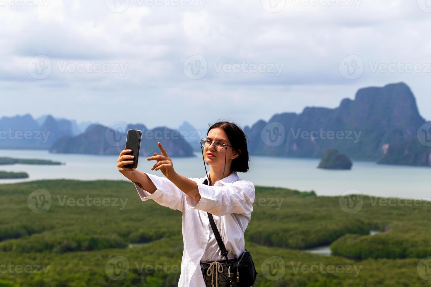 Young woman taking selfie with smartphone against stunning landscape of mountains and water, capturing moment of joy and adventure in nature's beauty. Female traveler is taking selfie with smartphone photo