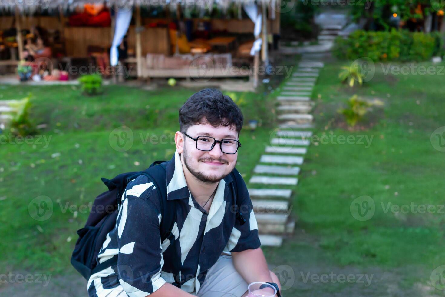 Traveler with backpack. Young man wearing stylish patterned shirt is sitting outdoors, surrounded vibrant greenery and stone pathway leading to cozy setting, creating relaxed and inviting atmosphere photo
