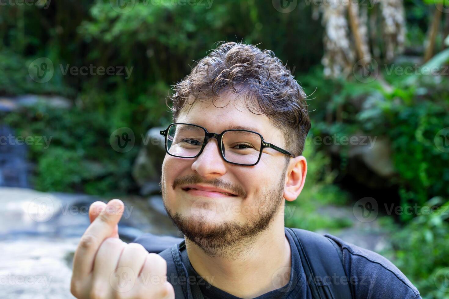 Young man with glasses is smiling and making heart gesture with his hands, surrounded by vibrant greenery and natural elements, creating joyful and friendly atmosphere. Traveler with backpack photo