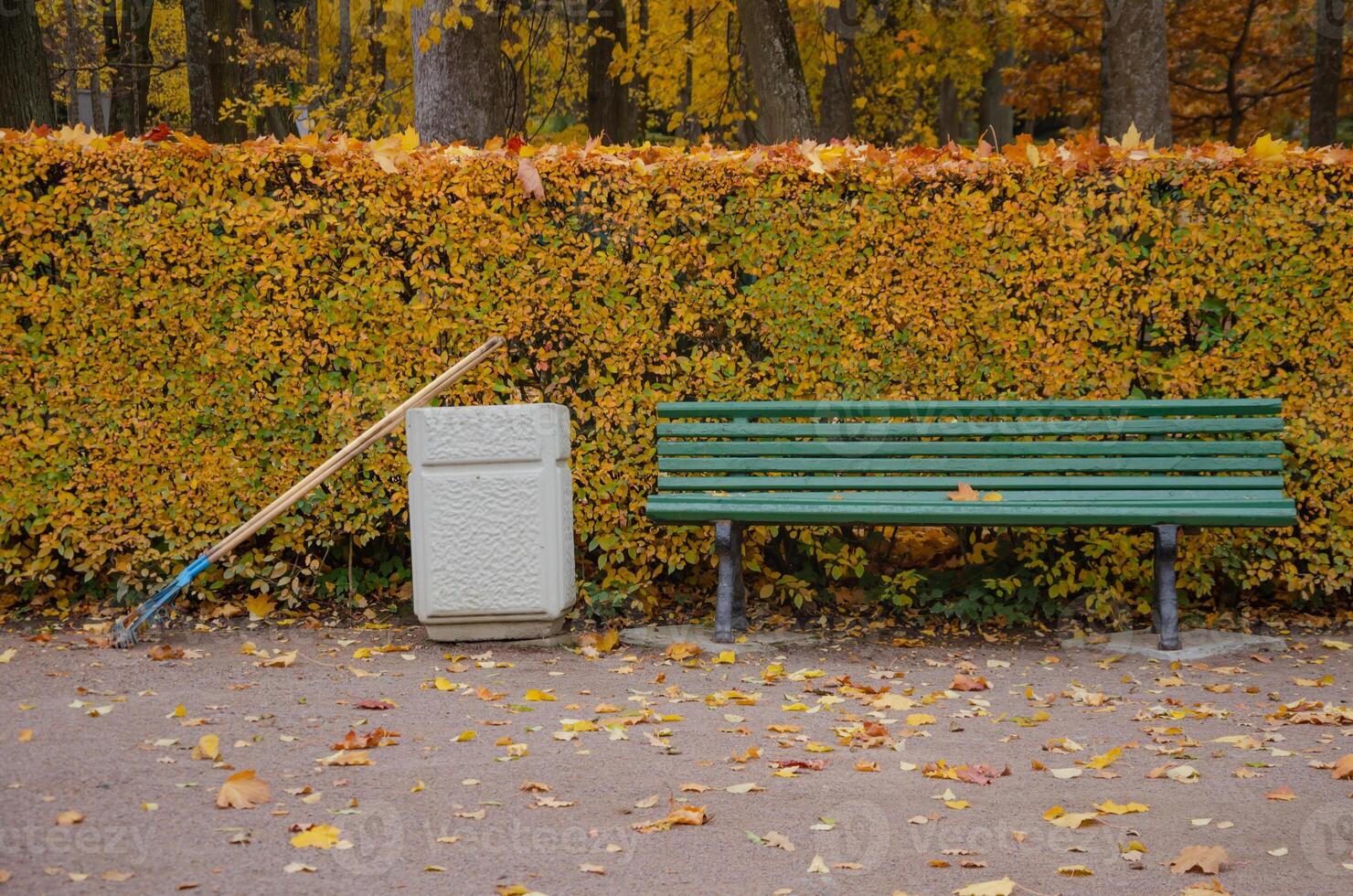In the autumn park, there is a bench, next to an urn and a rake for cleaning leaves. High quality photo