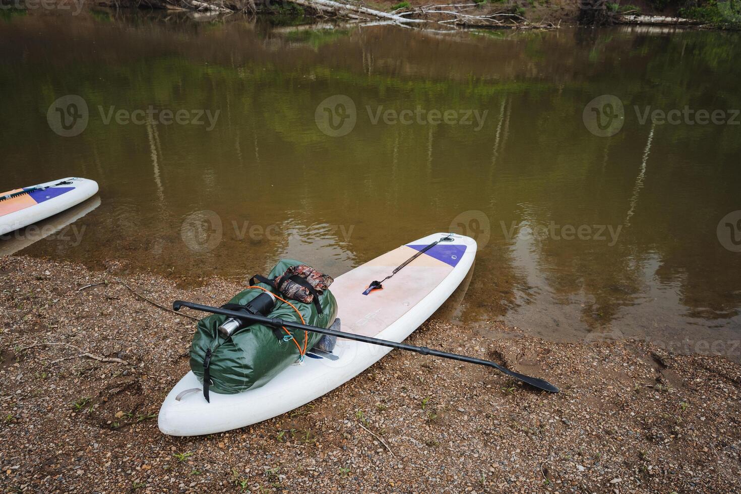 An exquisitely designed paddle board rests alongside a useful carrying bag on the soft sandy shore of a tranquil lake, sparkling brilliantly under the gentle, warm rays of the sun photo