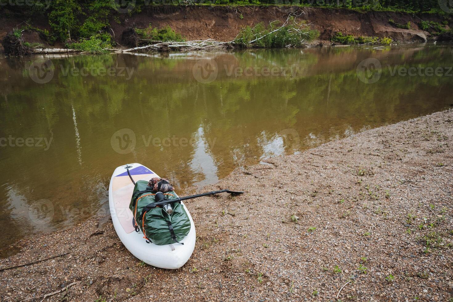 A colorful paddle board rests on a sandy river shore, waiting for someone to enjoy the tranquil waters, surrounded by lush nature and stunning views for an outdoor escape photo