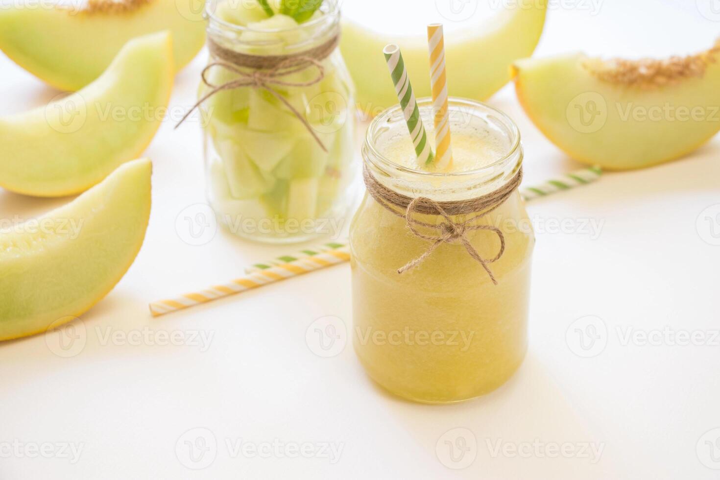 In front a jar with melon smoothie diagonally behind a jar with melon pieces near cut slices of melon, straws on a white background. Melon smoothie and salad in 2 jars. Horizontal. Daylight. photo