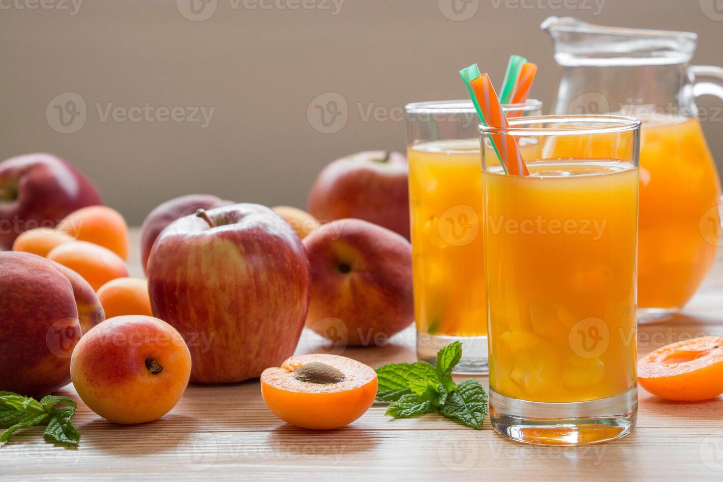 Apricot peach apple juice in glasses with ice near jar of juice and scattered apricots, peaches, apple mint on light wooden background. Apricot peach apple juice with ice. Horizontal. Daylight. photo