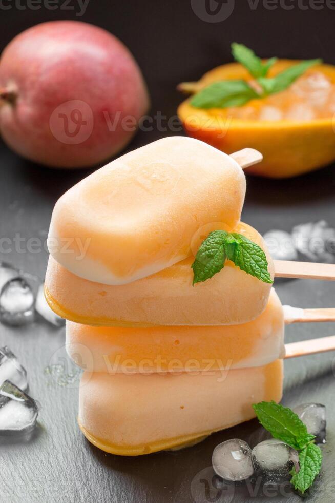 Column of mango sorbet sundae mix ice cream behind mango and cutted mango near ice cubes on a black background. Mango sorbet sundae mix ice cream popsicles. Vertical. photo