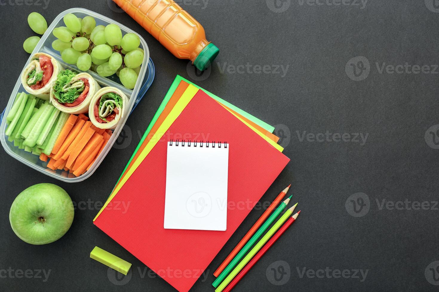 School supplies and lunch box with tasty rolls, cucumbers, carrots, grapes, apple, bottle of juice on black background, back to school concept with copy space. Flat lay. Top view. photo