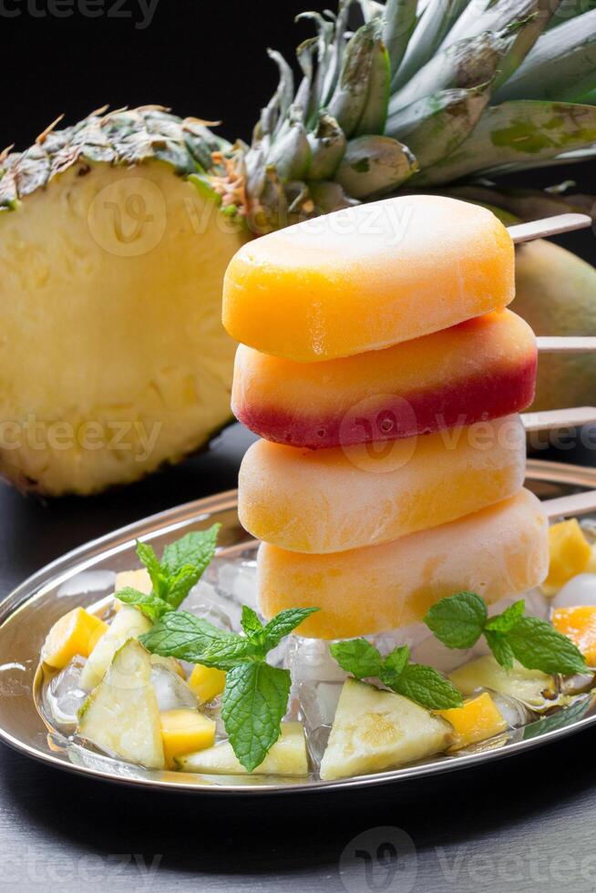 Column of sorbet ice cream popsicles from pineapple mango strawberry on silver plate with ice cubes and fruit pieces on a black background. Pineapple mango strawberry sorbet ice cream popsicles. photo