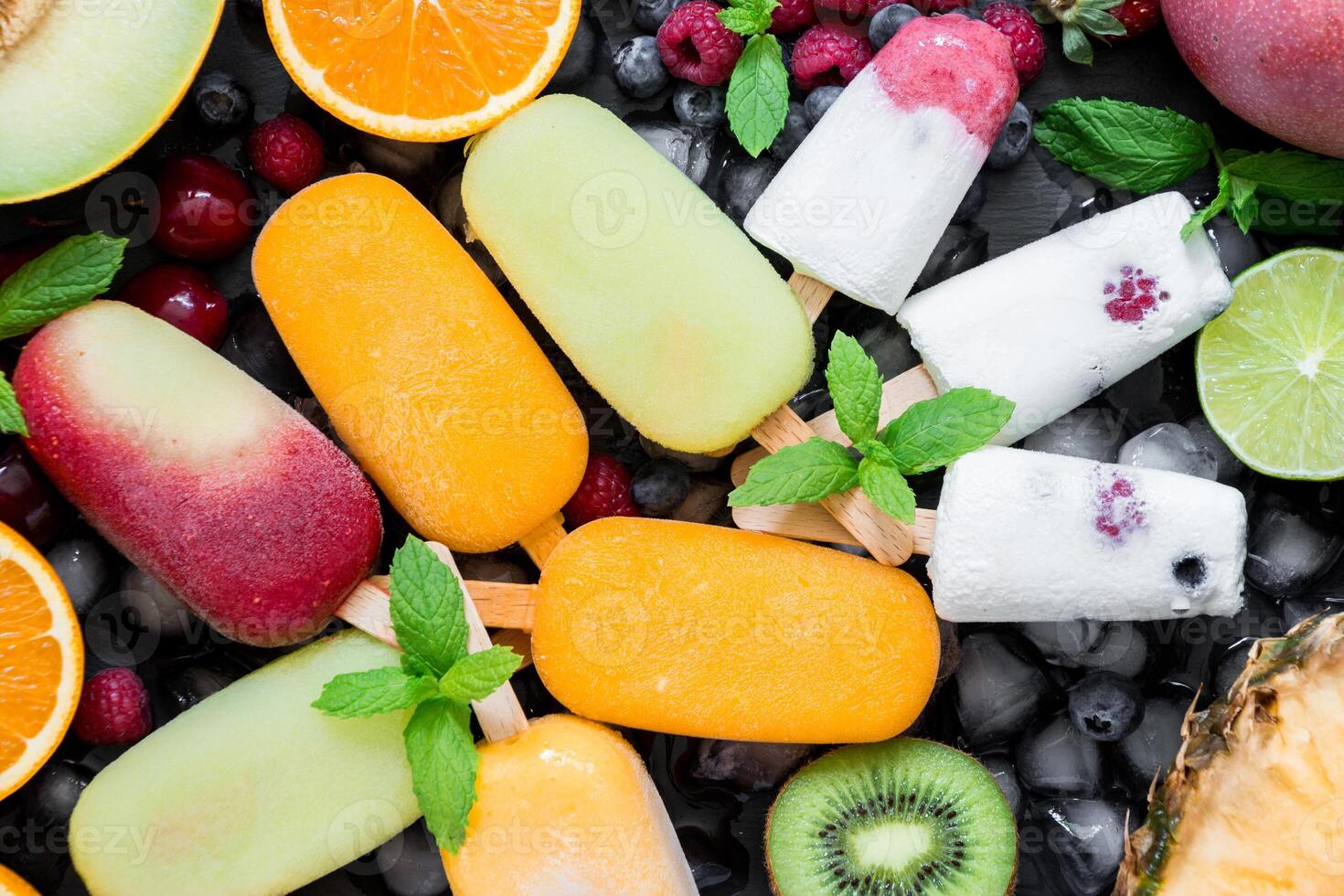 A vibrant array of berry flavored ice cream bars laid out flat, surrounded by slices of fresh fruits including melons, oranges, kiwis, and mint leaves for decoration. photo