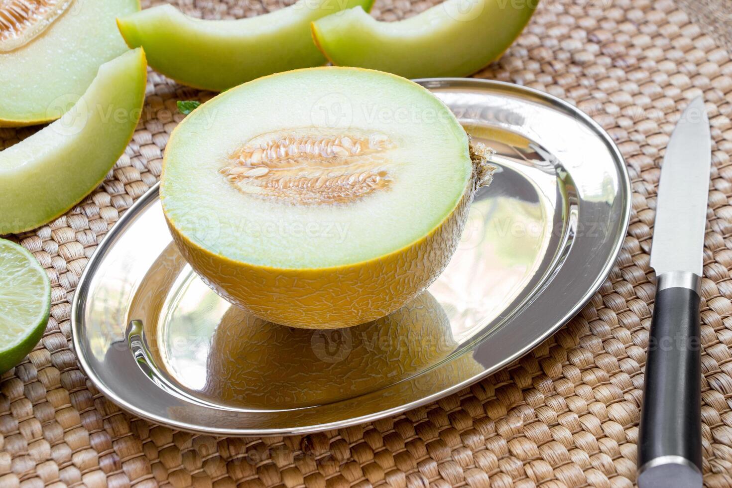 Diagonally at the center on a silver platter half of melon near melon slices, knife on a straw napkin background. Half of melon on silver platter. Horizontal. photo
