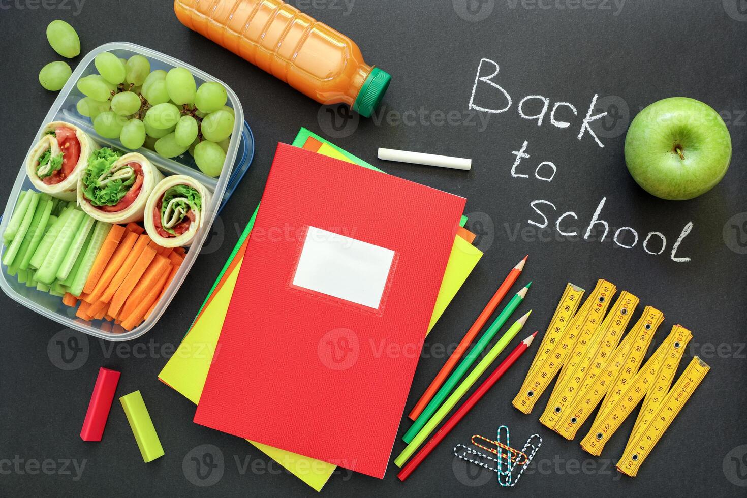 School supplies and lunch box with tasty rolls, cucumbers, carrots, grapes, apple, bottle of juice on black background, back to school concept. Top view. photo