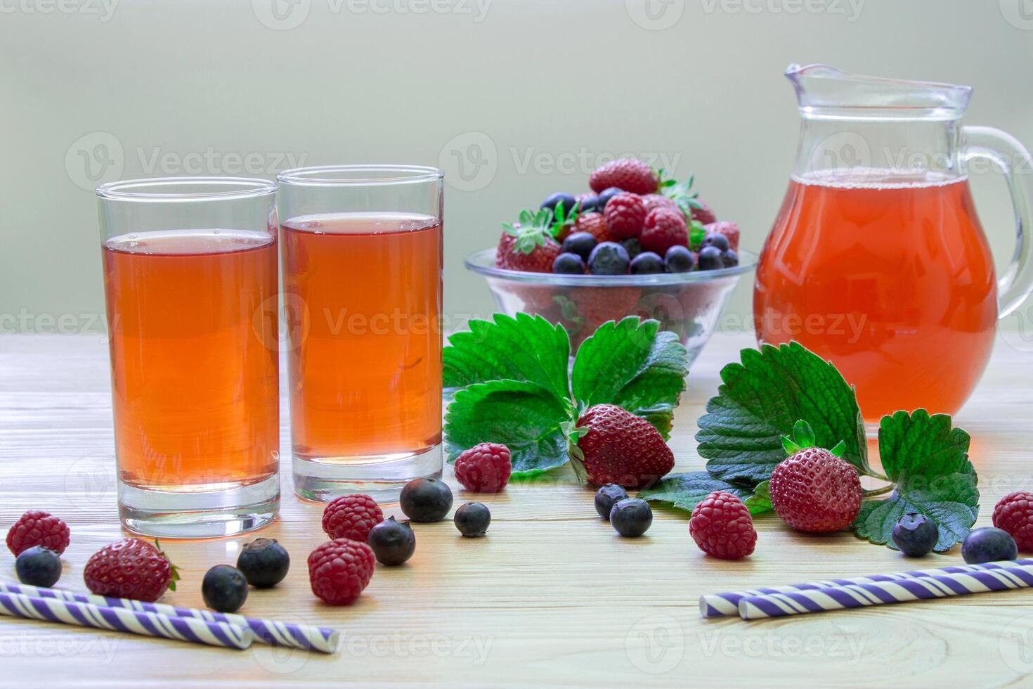 Left two glasses of berries compote, near bowl with berries, carafe compote on a light wooden background scattered berries, straws and empty space. Fresh berries compote and empty space. Horizontal. photo