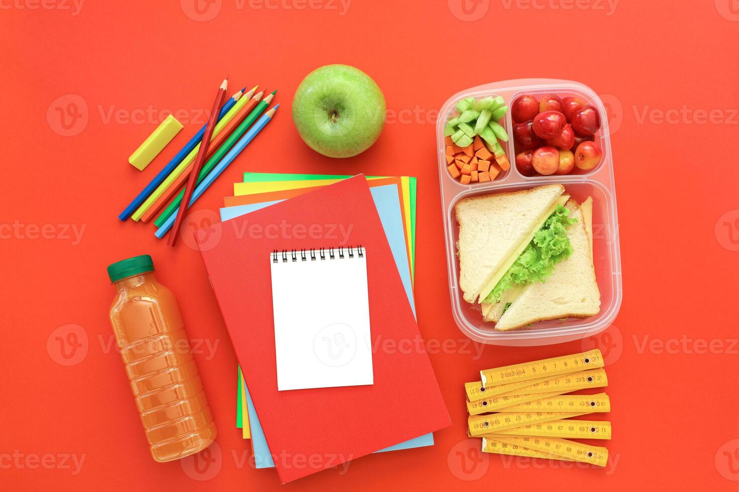 Colorful lunchbox filled with fruits, vegetables and sandwich sits beside green apple, school supplies, and drink on bright red surface. Ideal setting for preparing healthy meals. photo