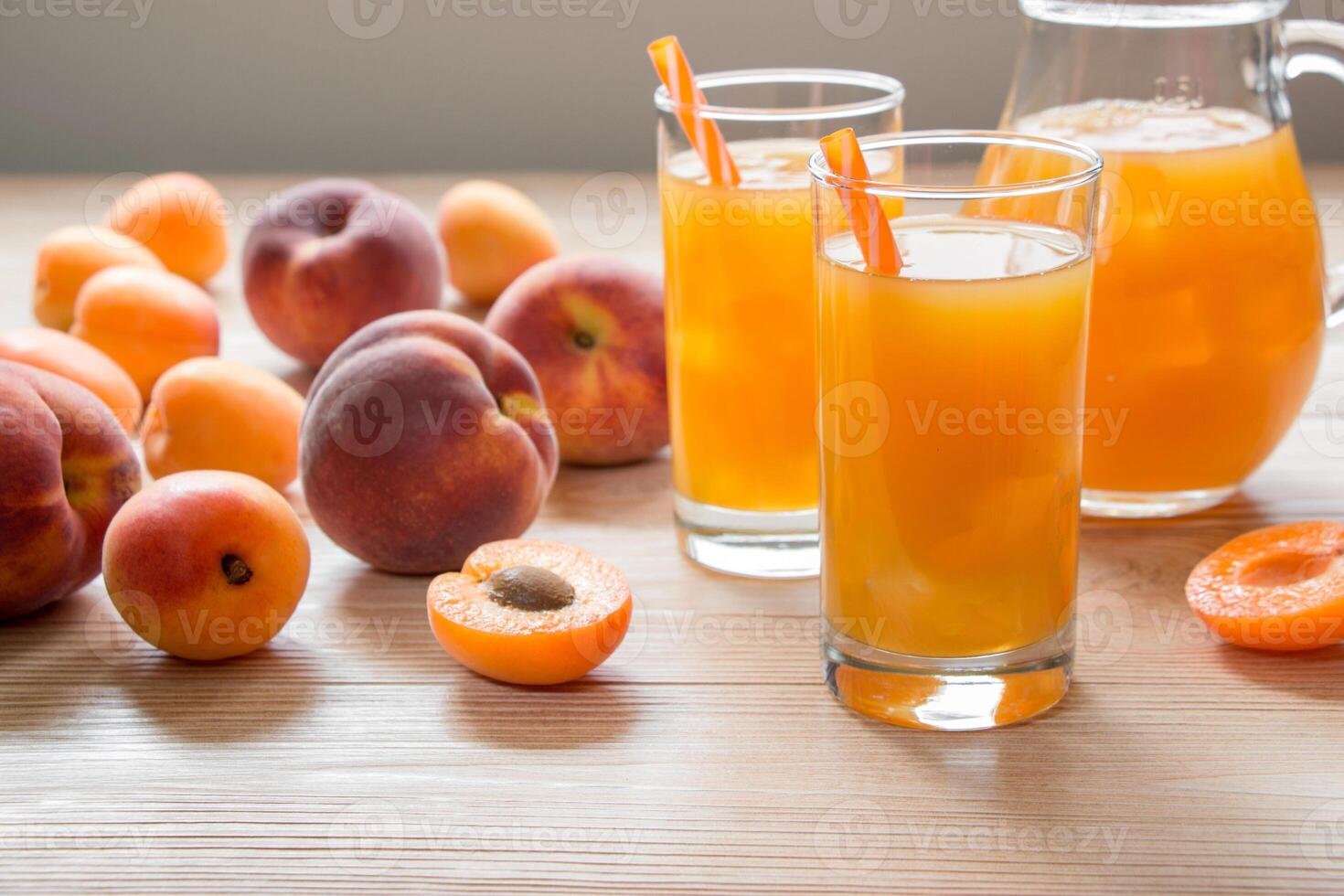 Glasses filled with bright apricot peach juice are placed on a wooden table, surrounded by fresh fruits and a pitcher. The warm sunlight illuminates the scene. photo