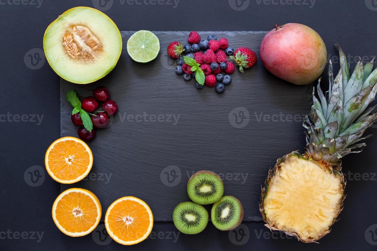 Rectangle from laid out fruit pineapple, mango, kiwi, oranges, lime, melon and berries cherries, strawberries, blueberries, raspberries in middle copy space on black background. Top view, flat lay photo