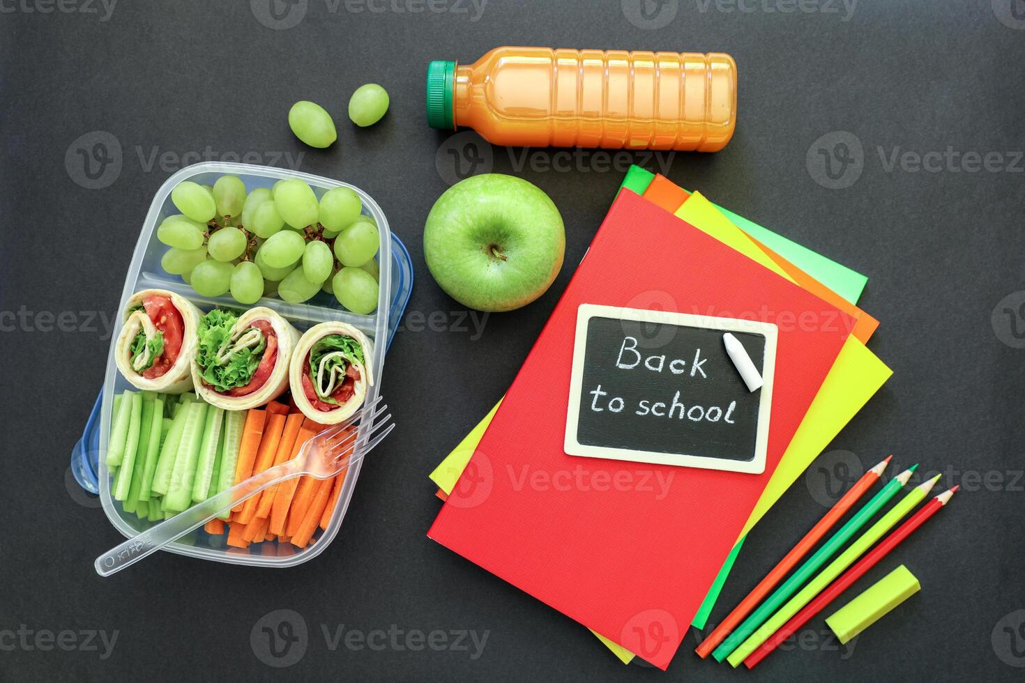 School supplies and lunch box with tasty rolls, cucumbers, carrots, grapes, apple, bottle of juice on black background, back to school concept. Flat lay photo