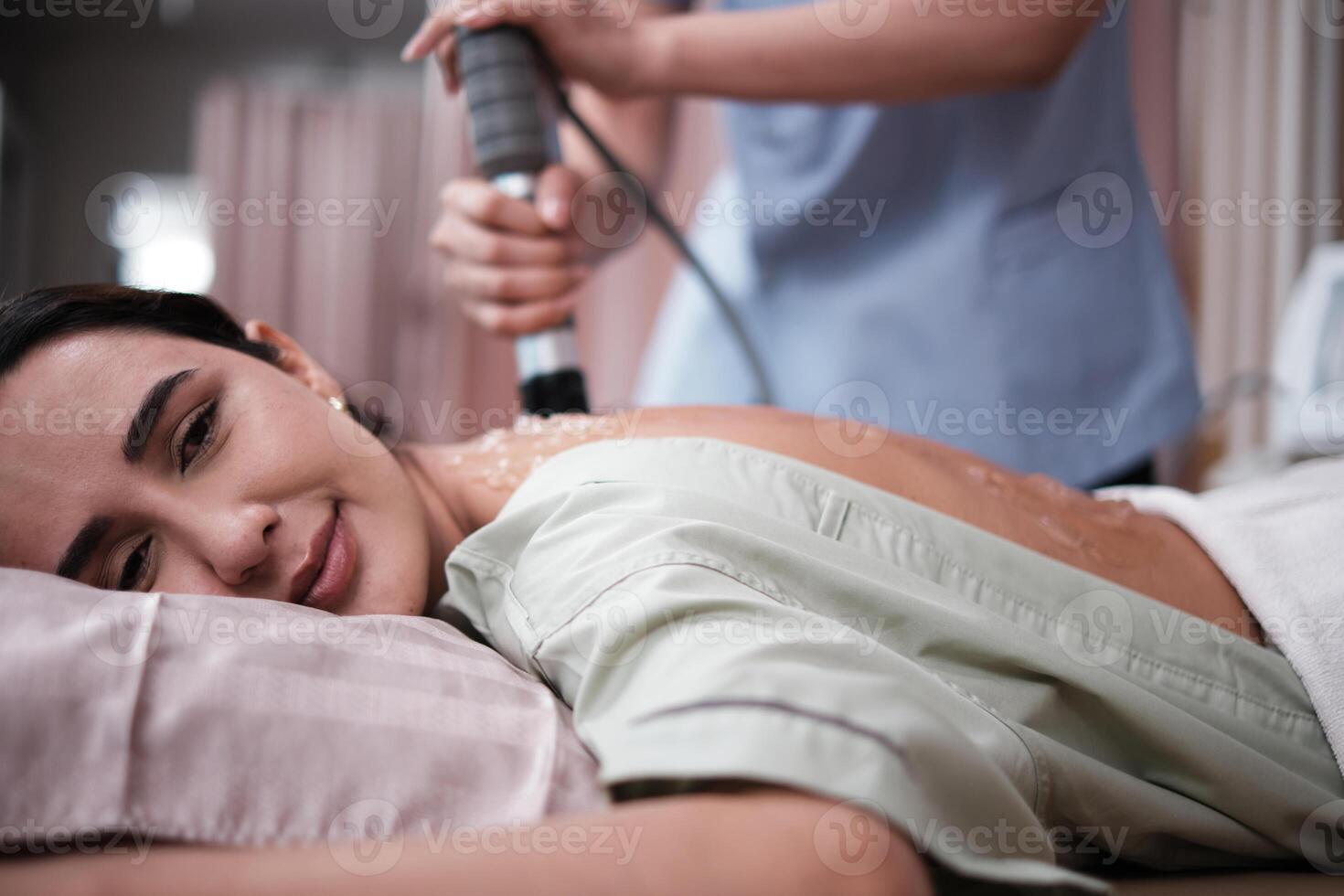 A Latino female patient recovered treated with ultrasound device for back pain relief procedure by Asian physical therapist, passive medical therapy at spa clinic, and wellness massaging healthcare. photo