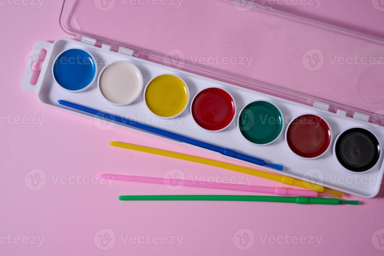 Set of watercolor paints in a plastic case with multi-colored brushes is located on a pink background. Top view photo