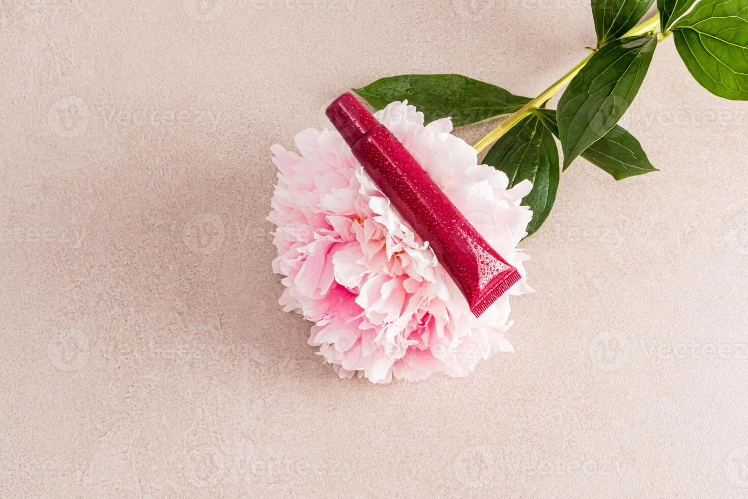 An unnamed tube of cosmetics lies on a large gorgeous pink peony flower. Beige stone background. A copy of the space. Blank tube layout photo