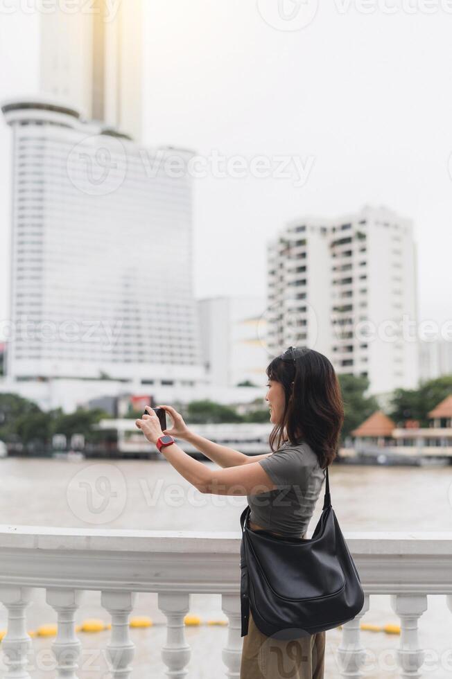 Solo world travel explorer southeast asian woman teenager blogger take photo with smartphone. Tourism concept to wait for boat Chao Phraya River express happy local cultural experience Bangkok