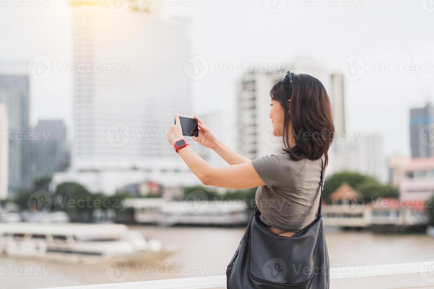 Happy solo Southeast Asian woman teenager blogger and travel explorer take photo with smartphone. Wait for express boat local cultural experience for Asia tourism Chao Phraya River Bangkok