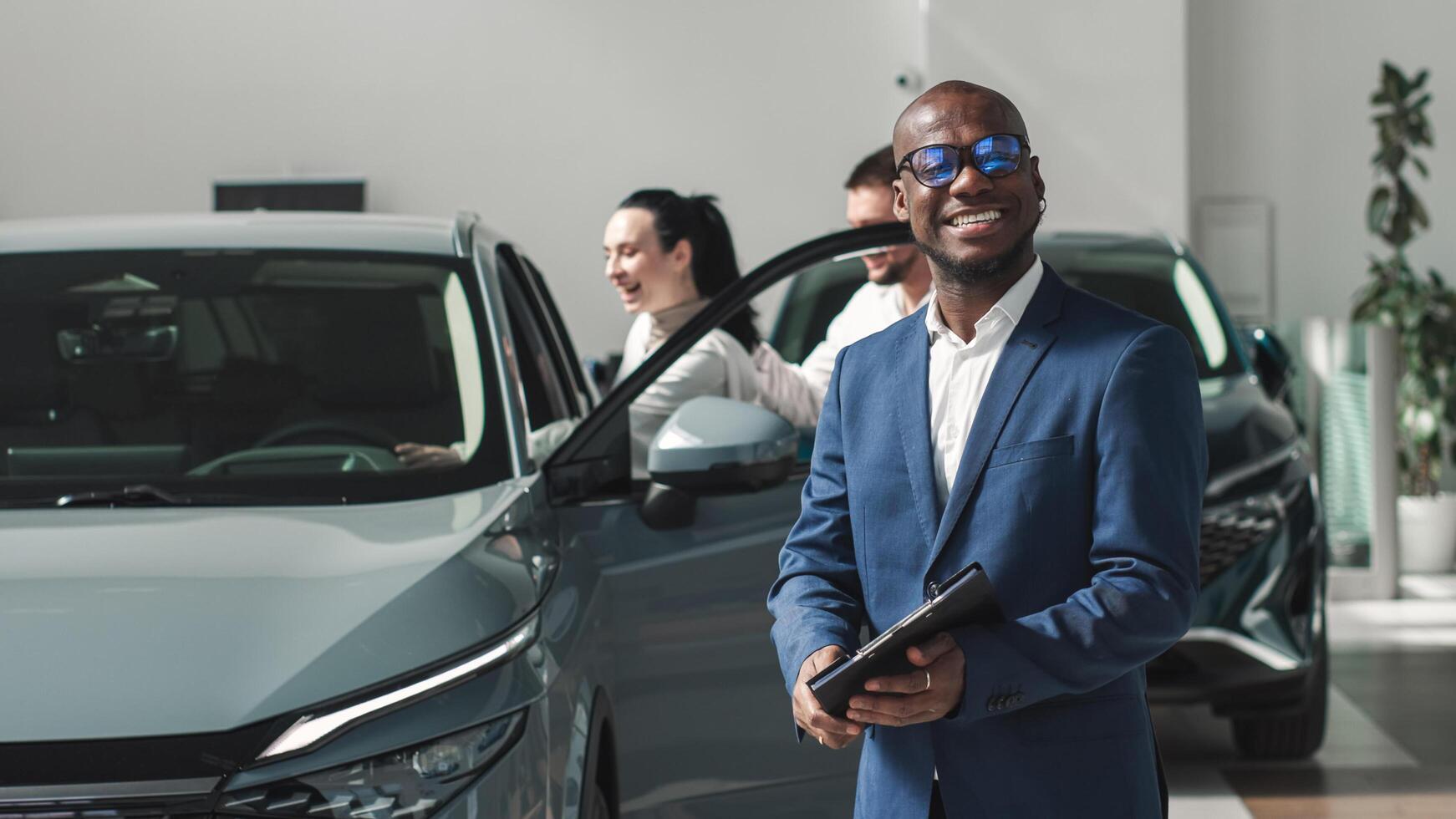 African-American dealer in the background of a happy couple after buying a new car in a modern car dealership. The concept of a long-awaited purchase, lease and test drive. photo