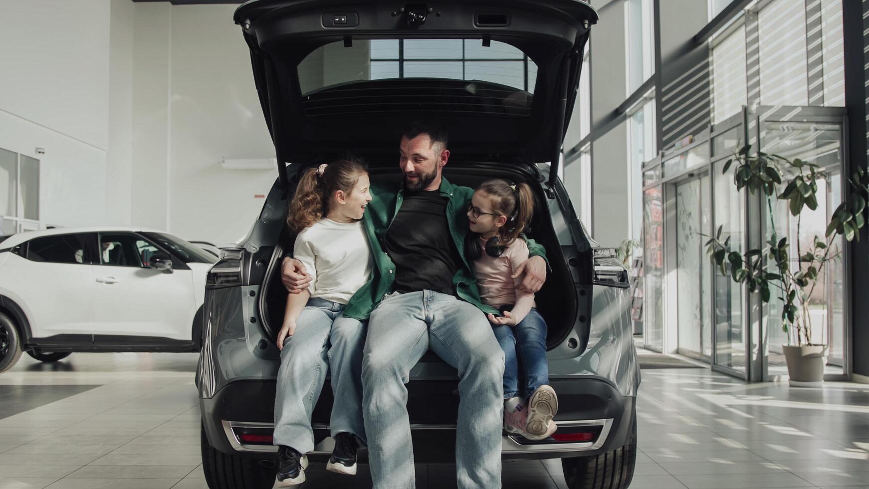 A happy father with his two daughters sits in the trunk of the car he just bought. The happy family is enjoying the purchase. The end of a test drive or purchase of a new, modern car photo