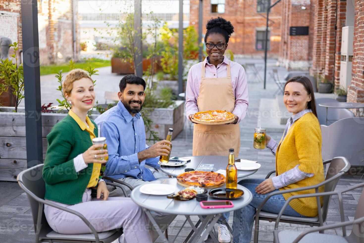 A diverse group enjoys pizza and drinks at an outdoor restaurant, served by a smiling waitress. The scene captures the joy of social gathering. photo