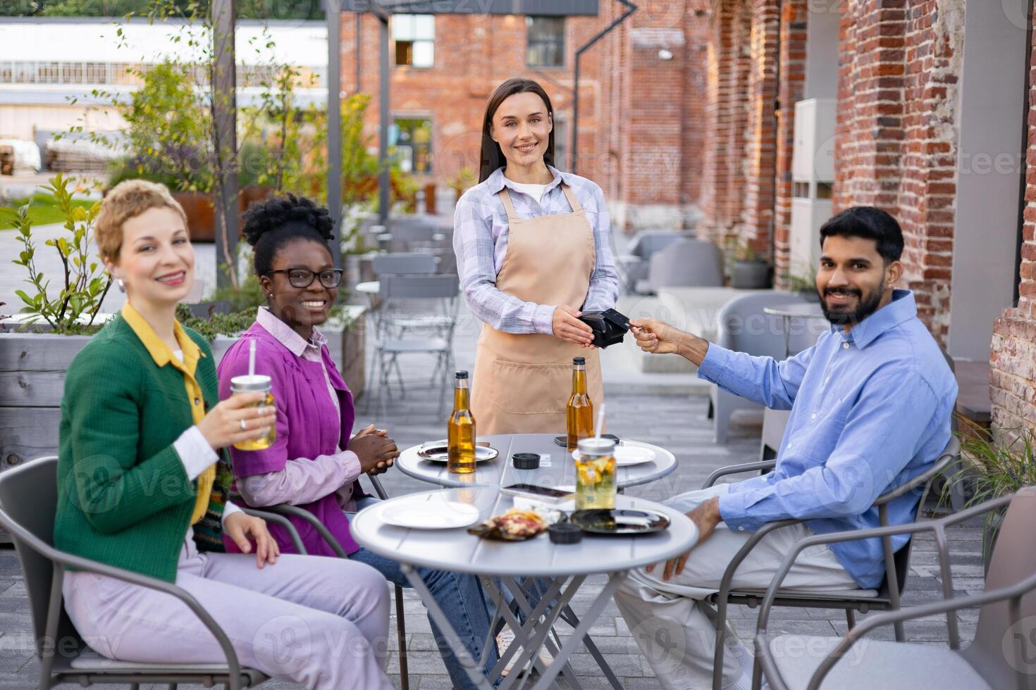 A female waiter in an apron serves customers at an outdoor restaurant, accepting payment via a portable terminal. photo