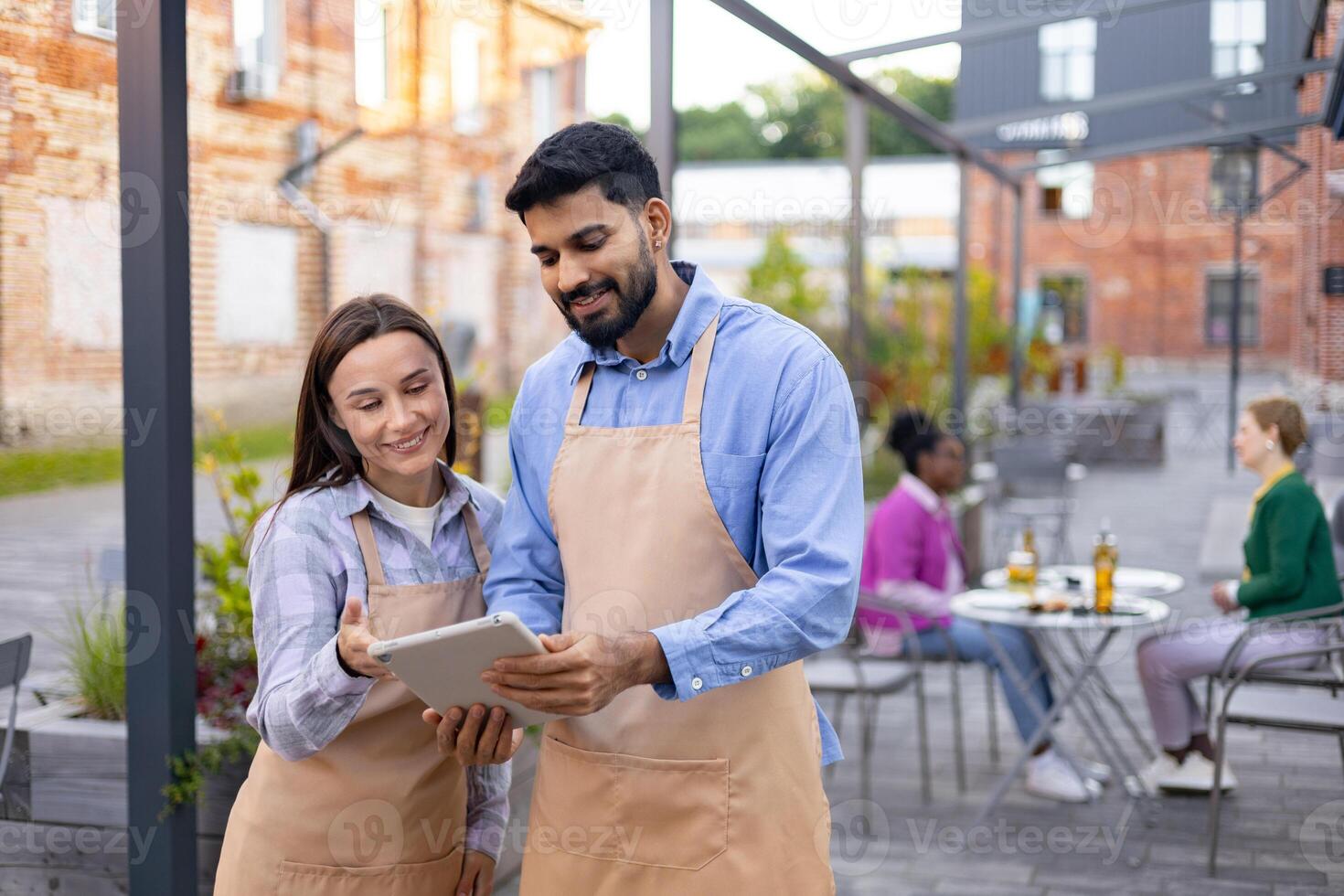 Two restaurant employees reviewing a tablet outdoors, smiling while discussing customer orders in a cafe setting. photo