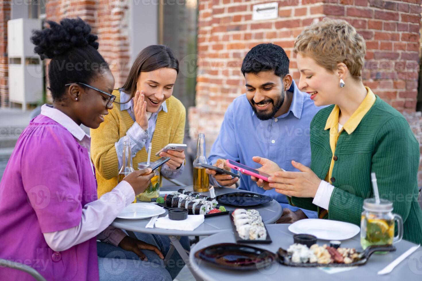 A diverse group of friends are gathered at an outdoor cafe, enjoying food and using their smartphones. photo
