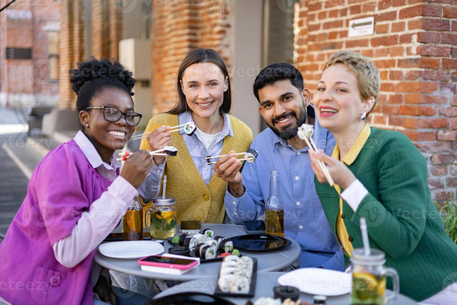 A diverse group of friends enjoys a sushi meal outdoors, smiling and using chopsticks at a table. photo